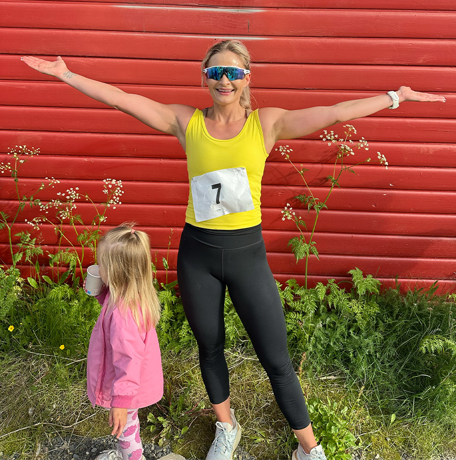 Kvinne i gul singlet med startnummer 7 står med utstrakte armer foran en rød vegg.