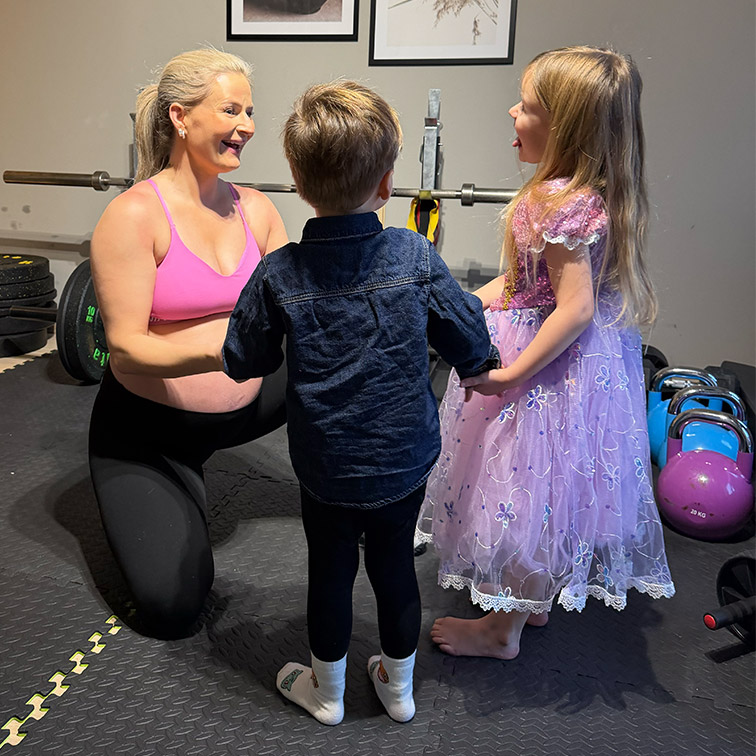 Gravid kvinne i treningsklær smiler til to barn i et treningsrom med vekter og kettlebells.